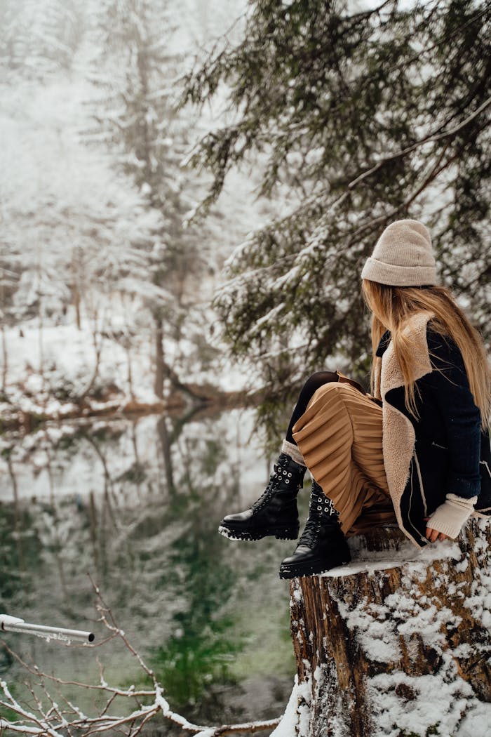A woman sitting on a tree stump by a serene winter lake in a snowy forest.